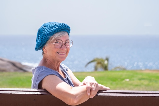 Mujer mayor sonriente con gorra azul sentada en un banco cerca del mar mirando a la cámara Anciana relajándose disfrutando de vacaciones o retiro bajo el sol Horizonte sobre el agua