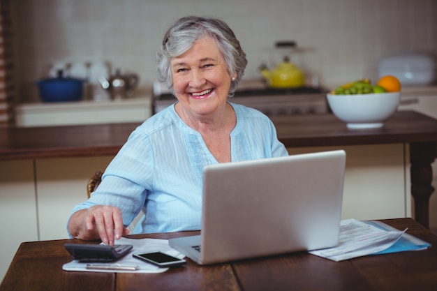 Mujer mayor sonriente que usa la calculadora y la computadora portátil