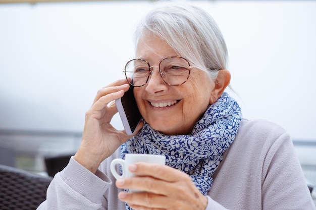 Una mujer mayor sonriente sentada al aire libre en una mesa de café hablando por teléfono inteligente sosteniendo un café espresso