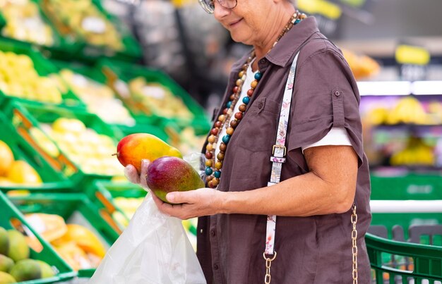 Mujer mayor sosteniendo dos mangos frescos en un primer plano de supermercado o tienda de comestibles Mujer sostiene dos mangos maduros con guantes protectores