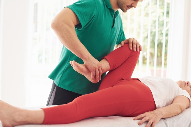 Una mujer mayor tiene una visita en la clínica acostada y rehabilitada Instructor masculino ayudándola