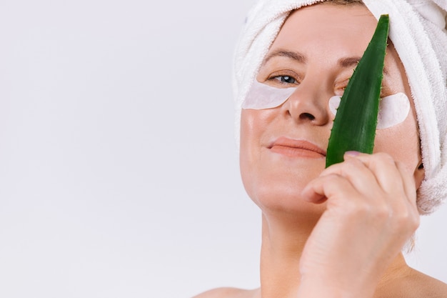 Mujer mayor con una toalla en la cabeza cubriéndose los ojos con una hoja de aloe vera sobre un fondo blanco con un espacio lateral vacío. Foto de alta calidad