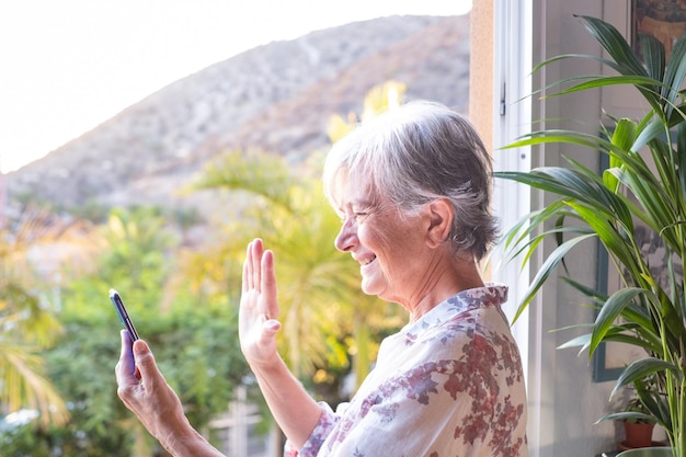 Mujer mayor en la ventana sosteniendo el teléfono móvil en videollamada con personas distantes. Sonriendo ancianos canosos disfrutando de la tecnología y las redes sociales