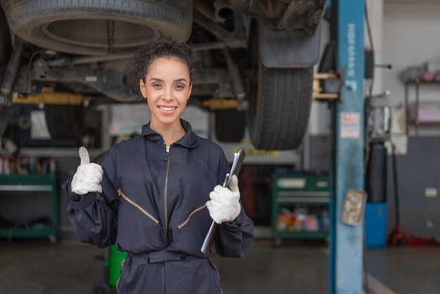 Mujer mecánico sonriendo mostrando los pulgares para arriba y trabajo de reparación de mantenimiento de un coche en el garaje de servicio automático