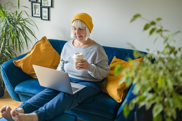 Mujer de mediana edad de 50 años sosteniendo café caliente trabajando en una laptop sentada en la oficina de casa