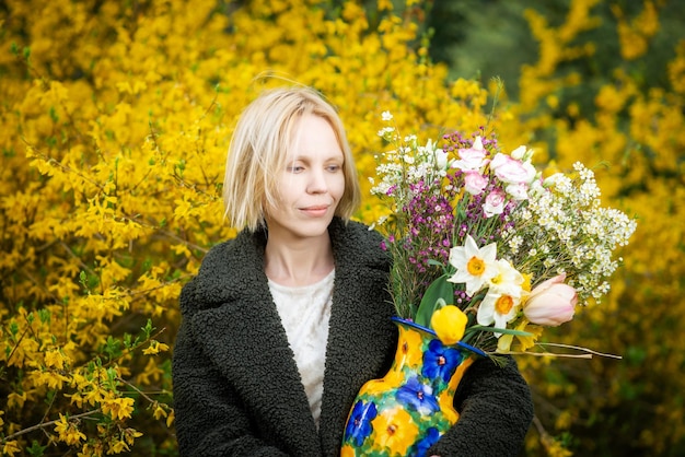 Una mujer de mediana edad con un abrigo verde con un ramo de varias flores sobre un fondo de flores amarillas florecientes