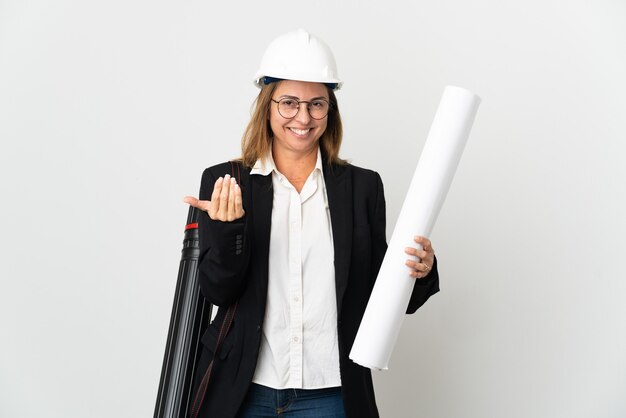 Mujer de mediana edad arquitecto con casco y sosteniendo planos sobre fondo aislado que invita a venir con la mano. Feliz de que hayas venido