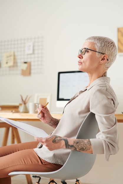 Mujer de mediana edad atractiva y pensativa con tatuajes analizando papeles en su propia oficina