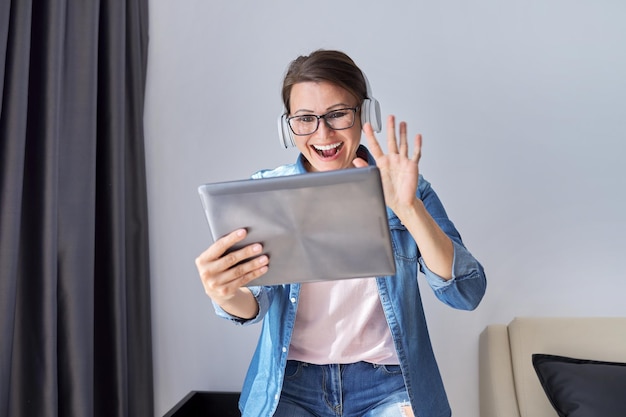 Mujer de mediana edad en auriculares con tableta digital en casa en sillón