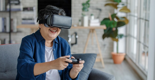 Mujer de mediana edad en auriculares VR sonriendo y jugando videojuegos mientras está sentado en el sofá el día de fin de semana en la sala de estar en casa