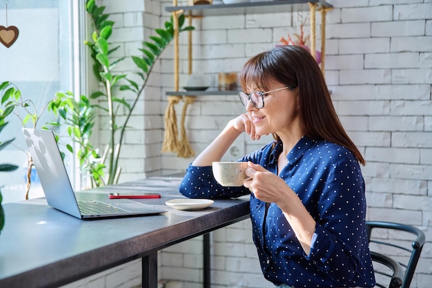 Foto mujer de mediana edad bebiendo café mirando la pantalla de su portátil sentada en una cafetería