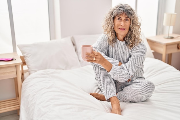 Mujer de mediana edad bebiendo una taza de café sentada en la cama en el dormitorio