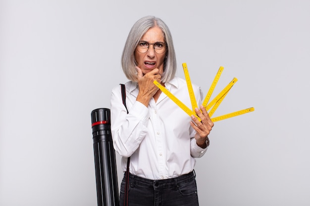 Mujer de mediana edad con la boca y los ojos bien abiertos y la mano en la barbilla, sintiéndose desagradablemente conmocionada, diciendo qué o guau. concepto de arquitecto