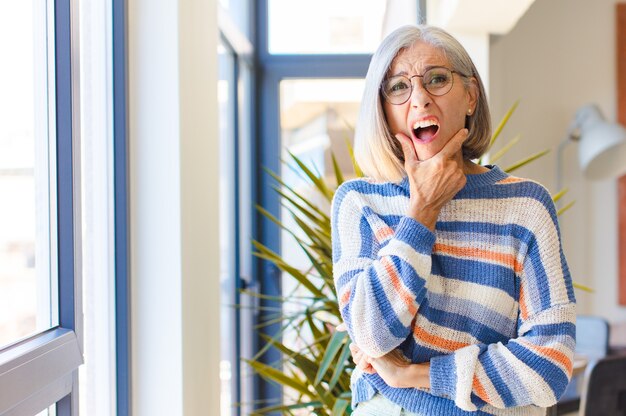 Mujer de mediana edad con la boca y los ojos bien abiertos y la mano en la barbilla, sintiéndose desagradablemente conmocionada, diciendo qué o guau