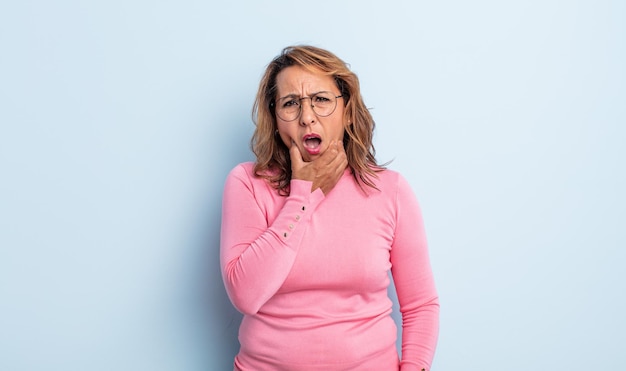 Mujer de mediana edad con la boca y los ojos bien abiertos y la mano en la barbilla, sintiéndose desagradablemente sorprendida, diciendo qué o wow