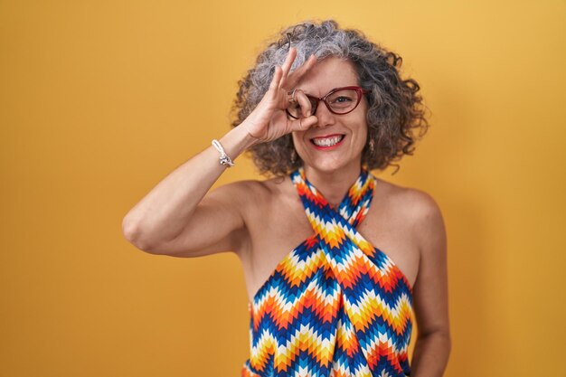 Mujer de mediana edad con cabello gris de pie sobre un fondo amarillo haciendo un gesto de ok con la mano sonriendo ojo mirando a través de los dedos con cara feliz