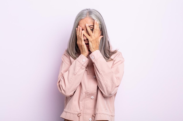 Mujer de mediana edad con cabello gris que se siente asustada o avergonzada, que mira a escondidas o espia con los ojos medio cubiertos con las manos