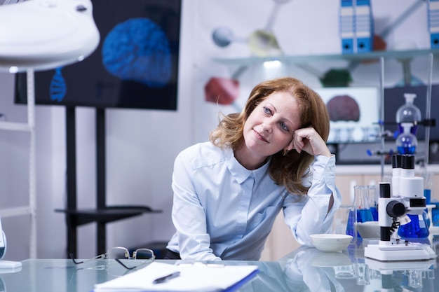 Mujer de mediana edad con cabello rojo en un laboratorio biomédico. Monitor con imagen cerebral de fondo.