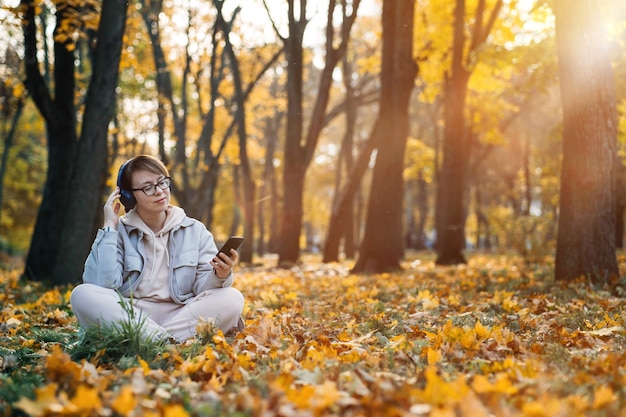 Mujer de mediana edad caucásica en auriculares escuchando música, aplicación de meditación en el teléfono inteligente y meditando en posición de loto en el parque de otoño. Aplicación de meditación, salud mental, autocuidado, concepto de atención plena.