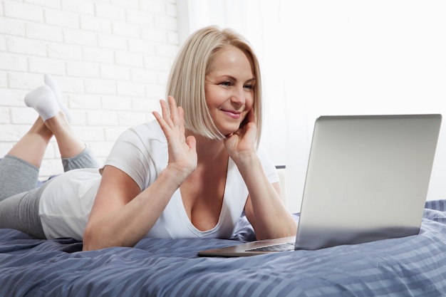 Mujer de mediana edad chateando en la cama usando una computadora portátil, navegando por internet, chateando.