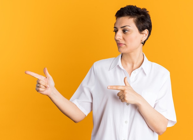 Mujer de mediana edad complacida mirando y apuntando al lado aislado en la pared naranja