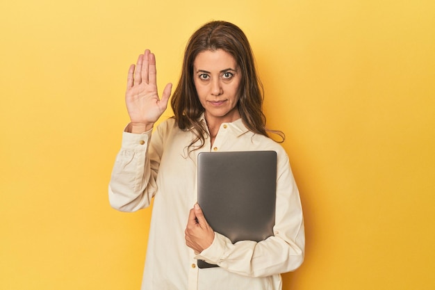 Mujer de mediana edad con una computadora portátil en amarillo de pie con la mano extendida mostrando una señal de parada