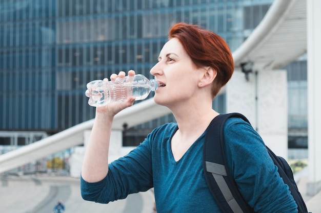 Una mujer de mediana edad con un corte de pelo corto bebe agua de una botella de plástico en el contexto de un moderno distrito de negocios urbano La necesidad de beber líquidos mientras hace calor