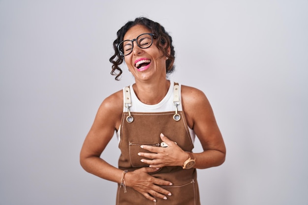 Foto mujer de mediana edad con delantal sobre fondo blanco sonriendo y riendo fuerte en voz alta porque es una broma divertida y loca con las manos en el cuerpo.