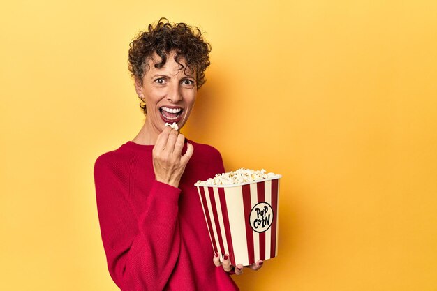 Mujer de mediana edad disfrutando de palomitas de maíz para una película sobre un fondo de estudio amarillo