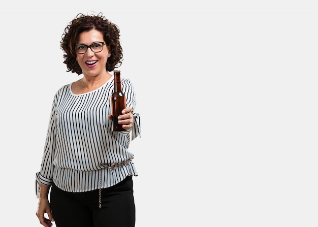 Mujer de mediana edad, feliz y divertida, sosteniendo una botella de cerveza, se siente bien después de un intenso día de trabajo, lista para ver un partido de fútbol en la televisión.