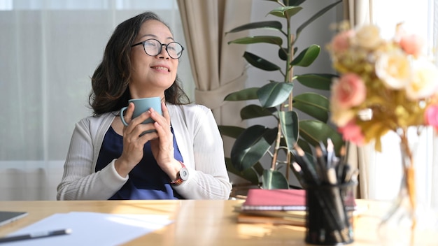 Una mujer de mediana edad feliz sosteniendo una taza de café disfruta de un estado de ánimo tranquilo y sin estrés en la sala de estar