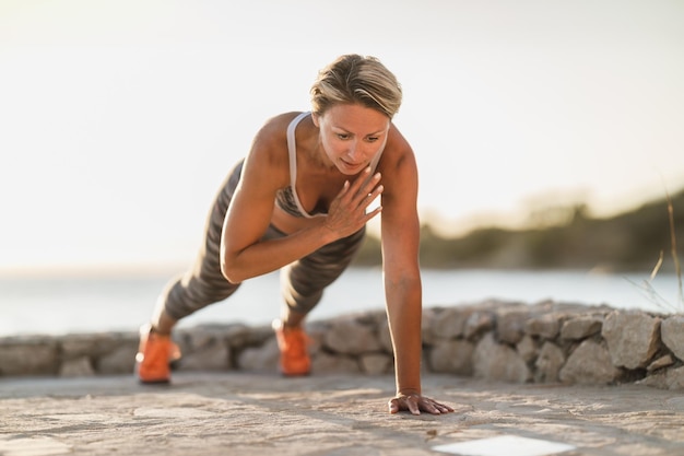 Foto mujer de mediana edad en forma haciendo ejercicios de tablones o flexiones durante un entrenamiento duro cerca de la playa del mar.