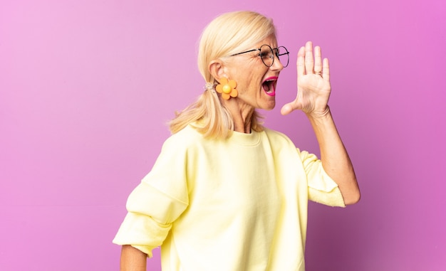 Mujer de mediana edad gritando fuerte y enojada para copiar el espacio en el costado, con la mano junto a la boca