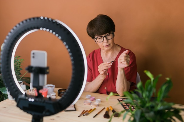 Mujer de mediana edad hablando de cosméticos con sombras de ojos de maquillaje y paleta de rubor y pinceles mientras graba su video Mujer madura haciendo video para su blog sobre cosméticos