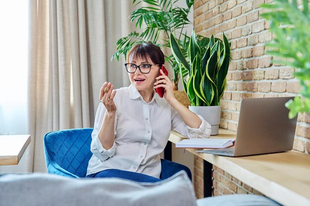 Foto mujer de mediana edad hablando por teléfono móvil sentada en el escritorio en casa con una computadora portátil