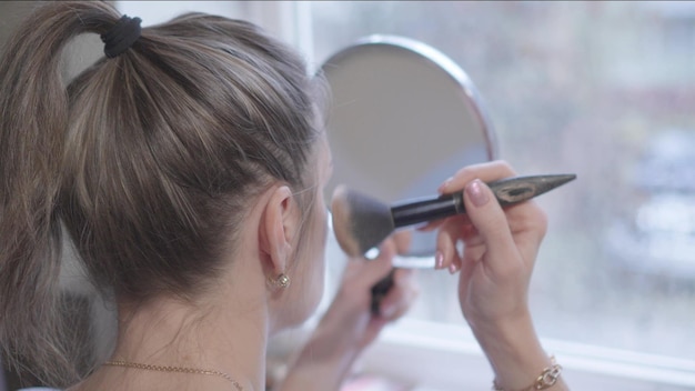 Foto mujer de mediana edad hace su maquillaje matutino cerca de la ventana mirándose en el espejo