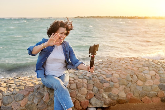 Mujer de mediana edad haciendo videollamadas en línea hablando riéndose del fondo del paisaje marino al atardecer