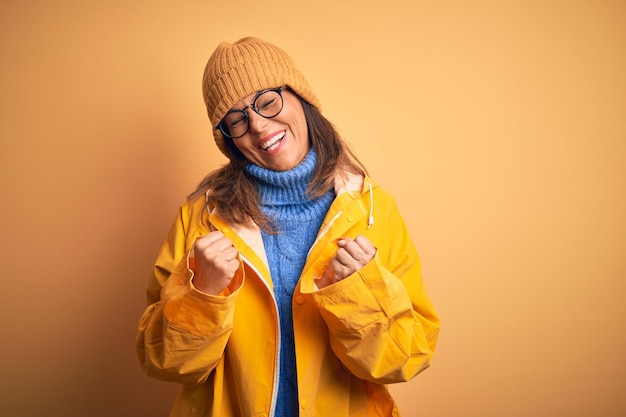 Foto mujer de mediana edad con impermeable amarillo y sombrero de invierno sobre un fondo aislado muy feliz y emocionada haciendo gesto de ganador con los brazos levantados sonriendo y gritando por el éxito concepto de celebración