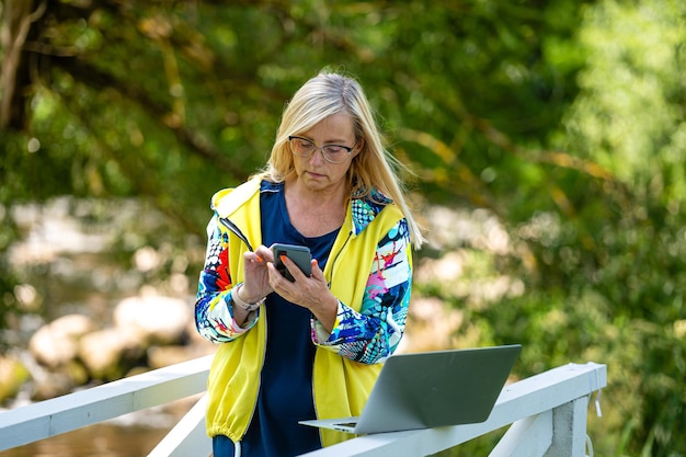 Mujer de mediana edad en el jardín trabajando desde casa usando una computadora portátil y hablando por teléfono