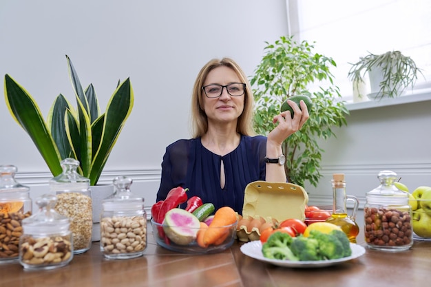 Mujer de mediana edad nutricionista profesional sentada a la mesa con verduras frutas huevos miasl nueces, mirando a la cámara, consultando, sosteniendo aguacate en la mano