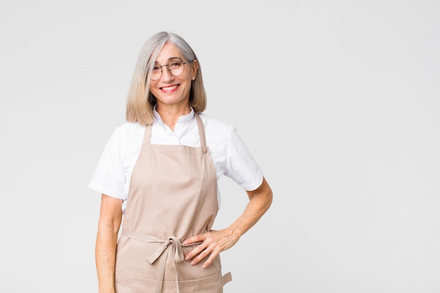 Mujer de mediana edad panadero sonriendo felizmente con una mano en la cadera y actitud segura, positiva, orgullosa y amigable en la pared plana