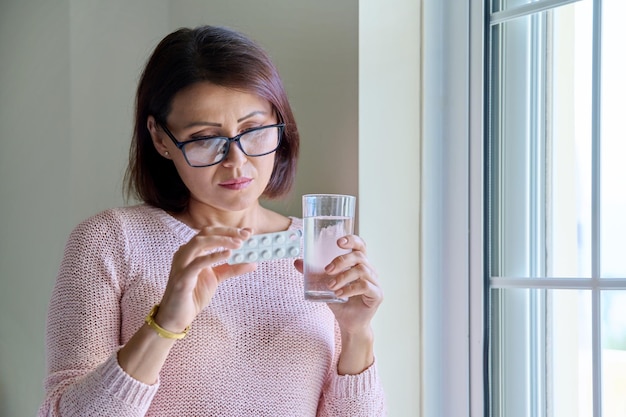 Mujer de mediana edad con pastillas y un vaso de agua en las manos en casa