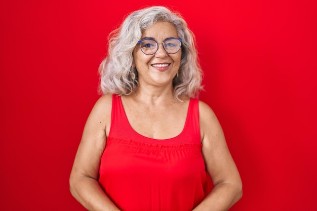 Mujer de mediana edad con el pelo gris sobre fondo rojo con las manos juntas y los dedos cruzados sonriendo relajada y alegre. exito y optimista