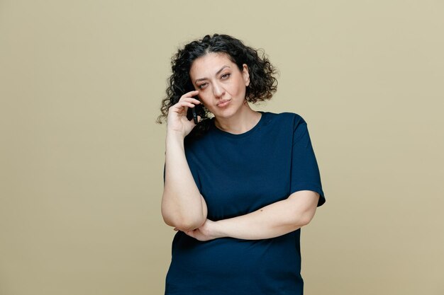 Mujer de mediana edad pensativa con camiseta manteniendo la mano bajo el codo mirando a la cámara hablando por teléfono aislada en un fondo verde oliva