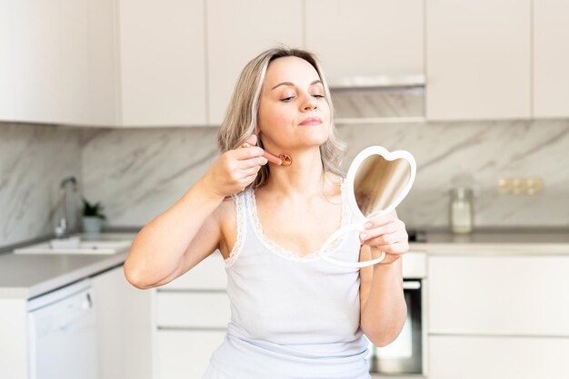 Mujer de mediana edad en pijama en la cocina con herramienta de masaje facial y espejo Procedimiento de belleza y antienvejecimiento para el cuidado de la piel Cuidado facial Cosmetología de la piel Rutina matutina Spa y bienestar en el hogar