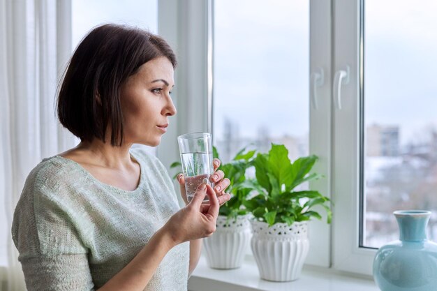 Mujer de mediana edad con píldora y vaso de agua en sus manos en casa en invierno