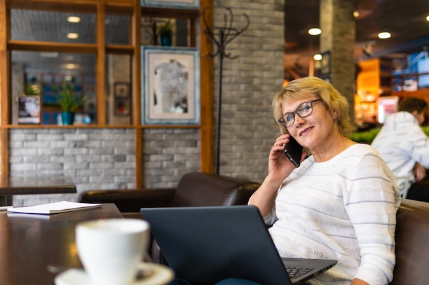 Una mujer de mediana edad con un portátil trabaja en un café de la oficina, es autónoma. Mujer con gafas sentado a la mesa con una taza