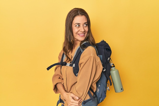 Mujer de mediana edad preparada para una caminata tiro de estudio amarillo mira a un lado sonriendo alegre y agradable
