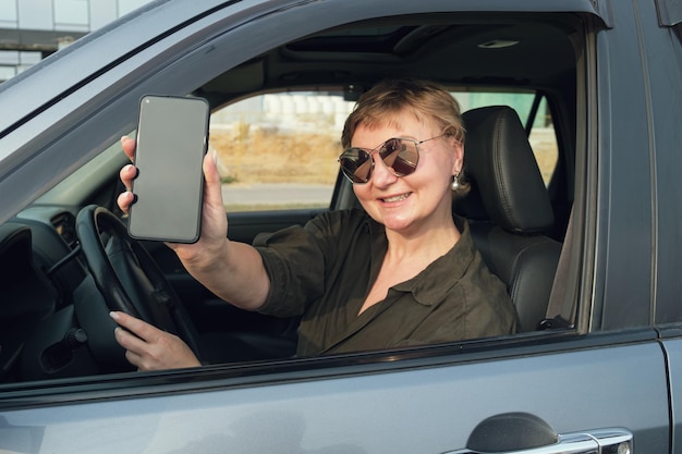 Mujer de mediana edad que conduce un automóvil sostiene su teléfono inteligente y sonríe