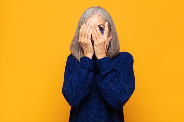 Mujer de mediana edad que cubre la cara con las manos, mirando entre los dedos con expresión de sorpresa y mirando hacia un lado